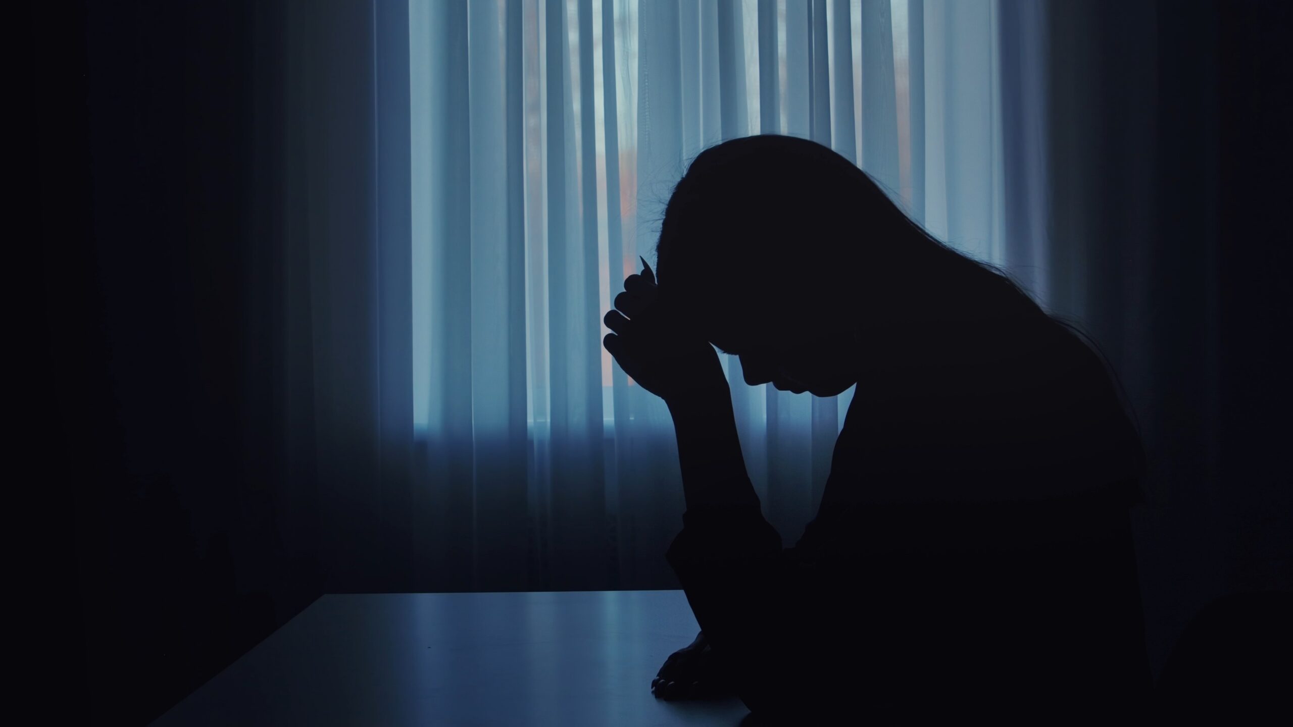 A silhouette of a woman with head down, seated alone in a dimly lit room