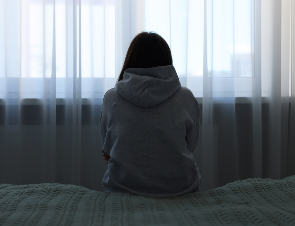 woman sits on bed in the dark, looking towards window.
