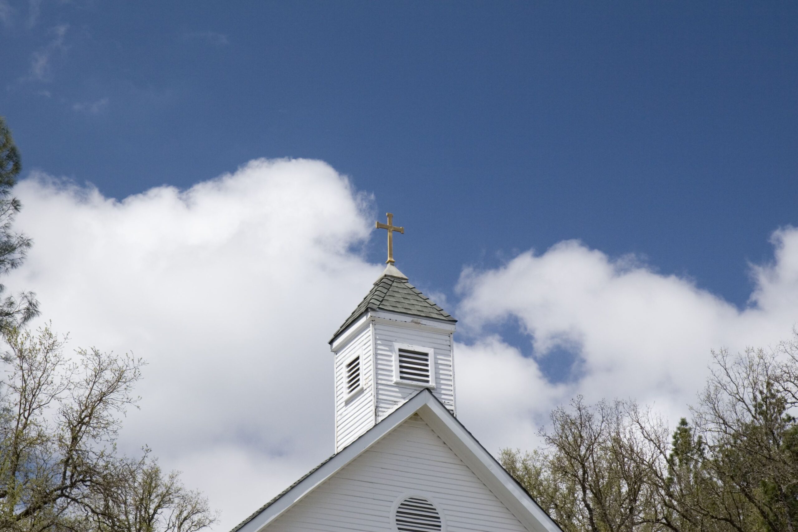 An older church steeple with brass cross against a partly cloudy sky